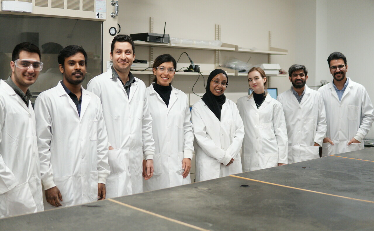 Dr. Mohajernia's 2024 research team. 8 people (4 men and 4 women) in white lab coats stand around a table/
