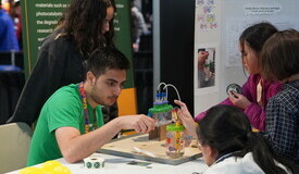 a man in a green shirt bends over a science demonstration, explaining it to three girls with dark hair